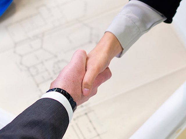 Stretta di mano tra due professionisti sopra i progetti di un edificio, con un casco di cantiere